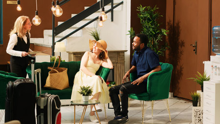 Hotel staff talking to guests in lobby, offering luxury services to people waiting in lounge area. Employee recommending tropical leisure activities to couple with room reservation.の写真素材