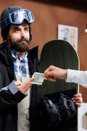 Detailed view of male snowboarder tipping employee with cash in ski hotel reception. Caucasian man in wintersport clothing giving gratuity to concierge at winter mountain resort.の写真素材