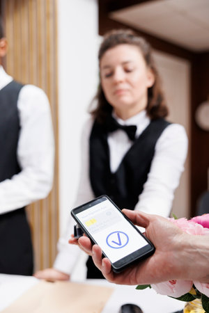 Female receptionist at front desk providing assistance to tourist using mobile phone for nfc...