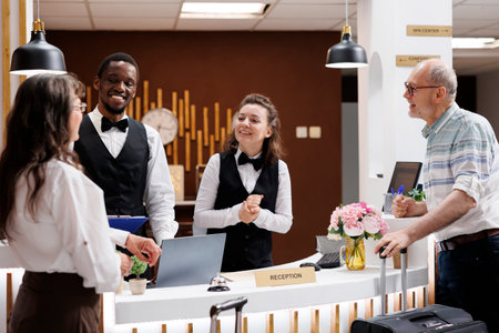 Two elderly tourists arrive with suitcases at hotel reception for check-in. Friendly smiling employees assists retired senior customers with booking and paperwork, creating welcoming atmosphere.の写真素材
