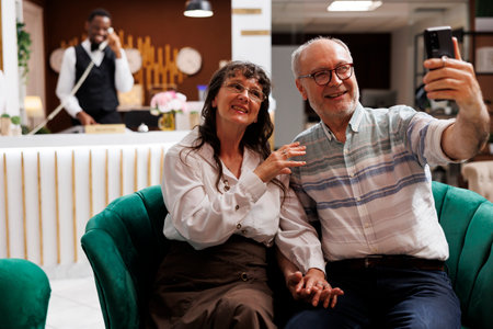 Elderly couple uses a mobile device to make a video chat while on vacation in a fancy hotel lobby. Retired senior travellers relax in the lounge area, talking with relatives on smartphone.の写真素材