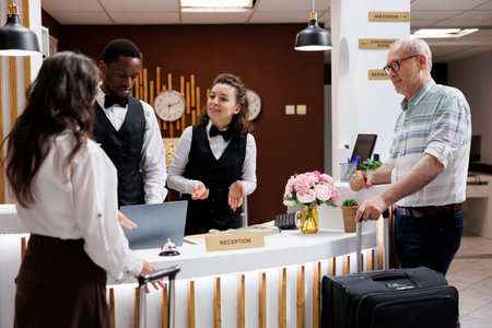 Assisting elderly guests at upscale hotel reception, concierge welcomes visitors with warmth and efficiency. Front desk staff ensures smooth check-in process for senior visitors in elegant lobby.の写真素材