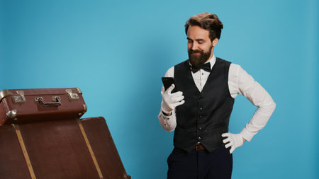 A hotel staff member in formal attire checks their smartphone while standing next to luggage against...