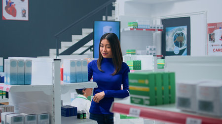 Smiling asian customer amidst pharmacy shelves, looking at medicine packages, trying to find suitable remedy for troubling ailments. Joyful cilent holding shopping bag, browsing through productsの写真素材