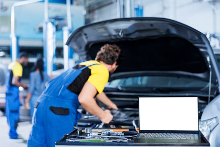 Mockup laptop on car service work station bench with technician in blurry background replacing engine. Isolated screen device next to qualified garage expert fixing client automobileの写真素材