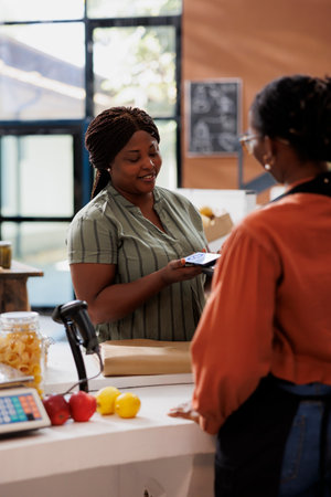An African American vendor smiles as a customer uses contactless payment at a bio food market. Freshly harvested, organic produce is available for sale.の写真素材