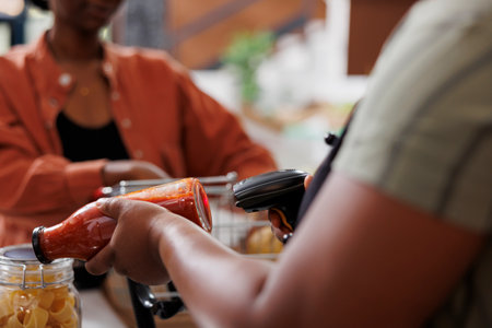 Closeup shot of african american cashier scanning bottle filled homemade pasta sauce for clients. Detailed view of barcode scanner being used by female vendor at checkout counter.の写真素材