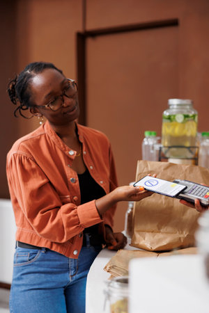 African American woman pays for her bio food using contactless payment on her mobile device. She holds a smartphone and supports cashless transactions at the locally grown market.の写真素材