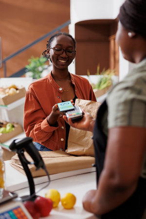 Female customer holding and using her mobile to device to make nfc payment for her sustainable goods. Young vendor holds out pos machine for client to pay with cell phone at loce eco friendly store.の写真素材
