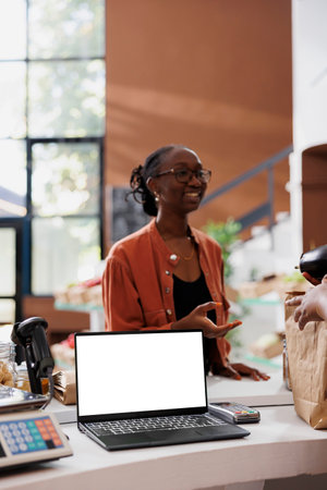 White screen visible on laptop while woman in glasses receives organic products. On counter is a standalone device with copy space, perfect for advertising an environmentally conscious bio food store.の写真素材