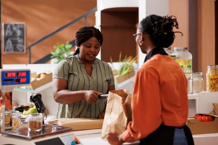 Female consumer paying with credit card at checkout counter, purchasing organic pantry staples at...