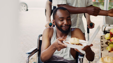 At outdoor food bank, close-up of poor male wheelchair user receiving free food. Volunteers serving warm meals and provisions to the handicapped and homeless people. Zoom-out, tripod shot.の写真素材