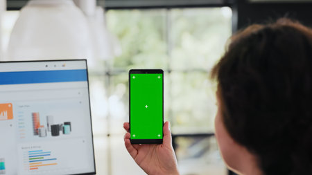 Worker examines phone with greenscreen sitting at workstation in business agency office, analyzing...
