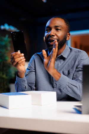 African american entertainer presenting mobile phone from brand sponsoring video, urging viewers to purchase it. Man does influencer marketing by advertising smartphone to audienceの写真素材