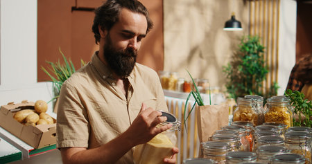Man in zero waste shop checking products before making purchase decision, making sure they are natural, dolly in shot. Client verifying pantry staples displayed in local neighborhood store are organicの写真素材