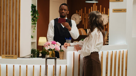 Hotel receptionist assists woman with check in, verifying identity on passport and retrieving reservation details. African american worker checks personal document of tourist at resort.の写真素材