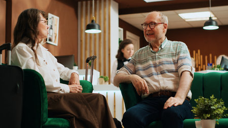 Retired people chatting in lounge area at five star resort, waiting to finish with registration forms for check in procedure. Senior couple on holiday talking about visiting landmarks. Handheld shot.の写真素材