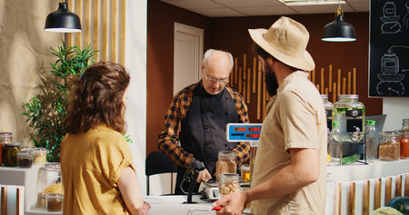 Older vendor assisting clients in zero waste shop with ethical sourced food items, inviting them at checkout. Couple in local store buying pantry staples bulk products, tracking shotの写真素材