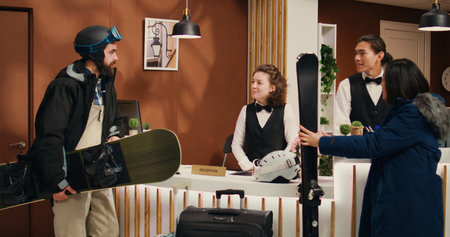 Tourists couple arrive at front desk, talking to receptionist about mountain slopes and skiing services included in reservation. Diverse employees greeting winter sport enthusiasts with gear.の写真素材