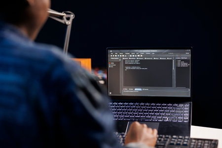 African American developer managing a system, typing on keyboard and laptop screen showing software...