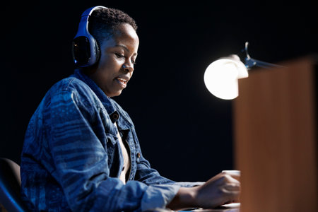 Young African American lady using a laptop in the apartment, browsing the internet, and listening to...