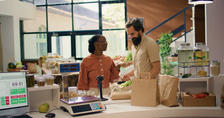 Seller assists customer in grocery store, recommending freshly harvested fruits and veggies on display. Couple searching for organic eco produce at local zero waste supermarket.の写真素材