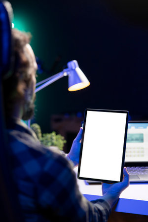 Over the shoulder view of a man grasping electronic tablet with blank chromakey mockup template. Young guy sitting at desk grasping digital device showing isolated white screen.の写真素材
