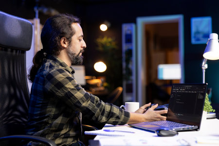 Male individual taking his wireless headset from table for music while working on his laptop. Bearded man touching his headphones while his device screen displays software codes.の写真素材