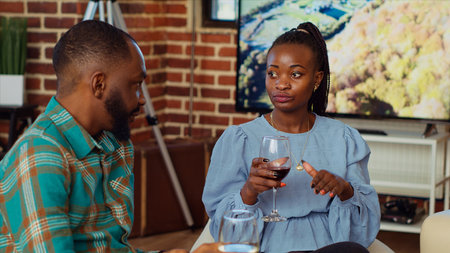 African american man at social gathering talking with his girlfriend on couch while enjoying glass of wine. Husband and wife guests at apartment party having pleasant conversation in brick wall homeの写真素材