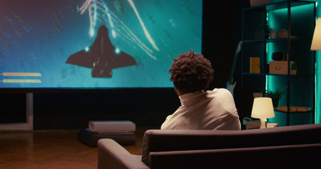 BIPOC man using cloud gaming service to play demanding SF videogame on widescreen TV. Panning shot...