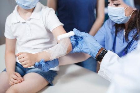 Close-up shows an injury on arm of patient being meticulously wrapped with sterile bandage by multicultural medical personnel. Photo focus on a youngster who is being treated by a doctor and a nurse.の写真素材
