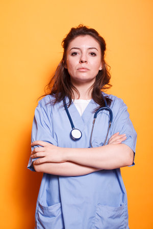 Medical practitioner wearing blue scrubs and standing against isolated orange background with her arms crossed. Portrait of female nurse with stethoscope and hands on chest, looking at camera.の写真素材