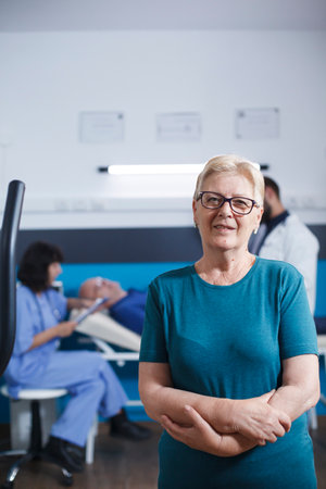 Old woman in portrait waits to work out for physical treatment with arms crossed. Senior patient with muscular injury gets ready for leg physiotherapy on stationary bike while she looks at camera.の写真素材