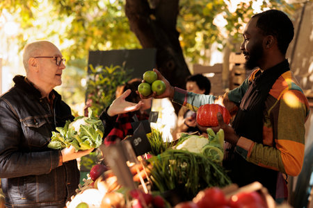 Senior man shopping for local organic produce at farmers market on weekend. Young African guy vendor...