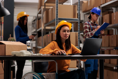 Young asian woman wheelchair user working on laptop in warehouse. Industrial storehouse employee...