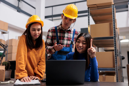 Asian delivery service managers analyzing logistics on laptop while standing near desk in warehouse. Storehouse package handlers and shipment operator looking at orders checklist on computerの写真素材