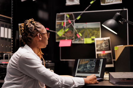 Investigator analyzing evidence board and checking photos on laptop. African american woman lawyer preparing for case while working late in police detective office room at nightの写真素材