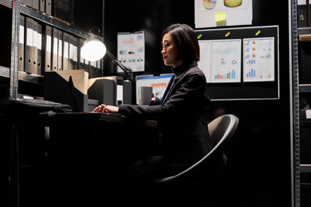 Business invoicing employee woman imputing accountancy market data analysis paperwork on laptop with cup of coffee in hand. Archival depository with folders in cabinet bookshelves and flowchartsの写真素材