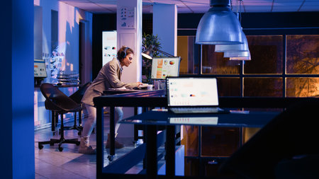Happy employee analyzing graphs wearing headset, working at night to increase business profit....