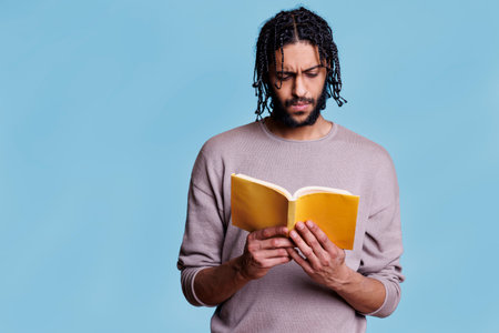 Concentrated arab man reading paperback book with yellow cover. Focused young person with thoughtful expression studying literature, holding softcover textbook on blue backgroundの写真素材