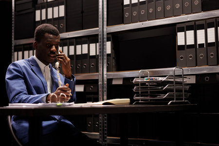 African american businessman discussing management research with remote employee using landline phone. Manager working late at night with administrative files in corporate depositoryの写真素材