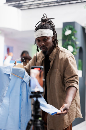 African american influencer promoting fashion store on social media, filming live video on mobile phone. Blogger showing male formal shirt on smartphone camera to advertise new collectionの写真素材