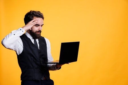 Happy modern waiter using laptop in studio and working at five star restaurant, searching for new menu online on own webpage. Professional employee looking to develop business with pc.の写真素材