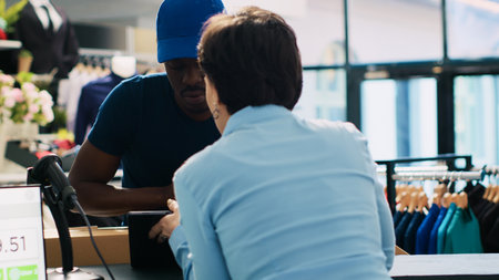 African american courier holding pile with carton boxes, discussing shipping details with store manager in modern boutique. Deliveryman signing shipment report on tablet computer. Fashion conceptの写真素材