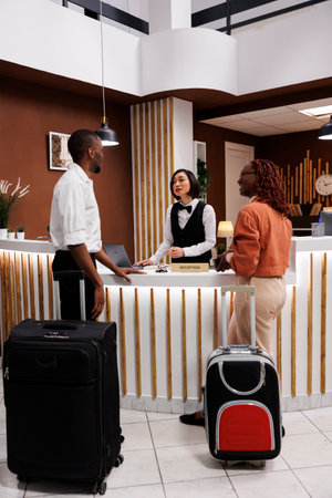 Asian woman welcoming couple at hotel, reviewing guests information before doing check in process to show room. Receptionist registering people arriving at resort, front desk.の写真素材