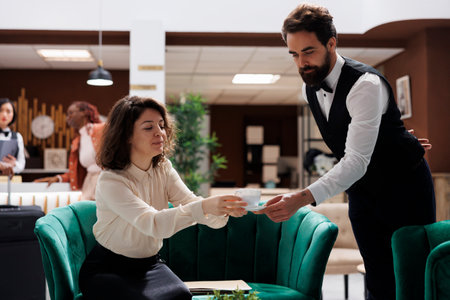 Hotel staff serving manager with coffee, giving drink to hotel supervisor in lobby space. Young administrator sitting in lounge area, receiving beverage from employee with professionalism.の写真素材