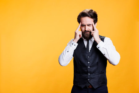Skilled waiter deals with headache against yellow background in studio, feeling sick with pain and rubbing temples. Young man being overworked with bad migraine, dealing with burnout.の写真素材