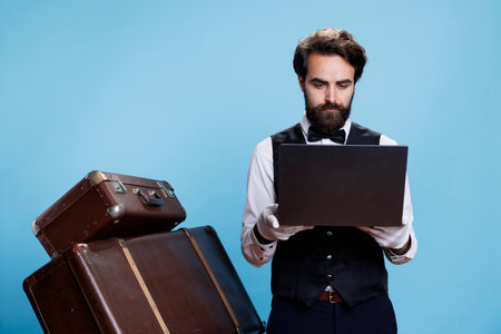 Hotel porter in suit using laptop against blue background, showcases professional hotel expertise. Classy doorkeeper with gloves and tie checking online bookings, hospitality industry.の写真素材