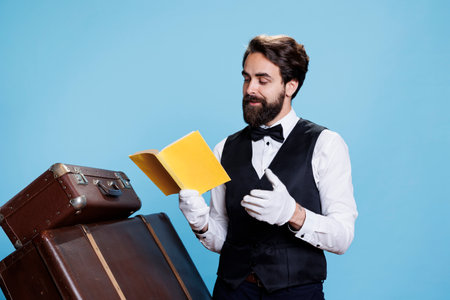 Stylish employee enjoys fiction book while on duty, taking care of trolley bags in studio. Male doorkeeper reading literature pages in front of camera, having fun with classic hobby.の写真素材