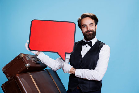 Hotel worker showing ad with speech bubble, holding empty isolated red billboard icon. Elegant doorkeeper presents blank copyspace cardboard sign on camera against blue background.の写真素材